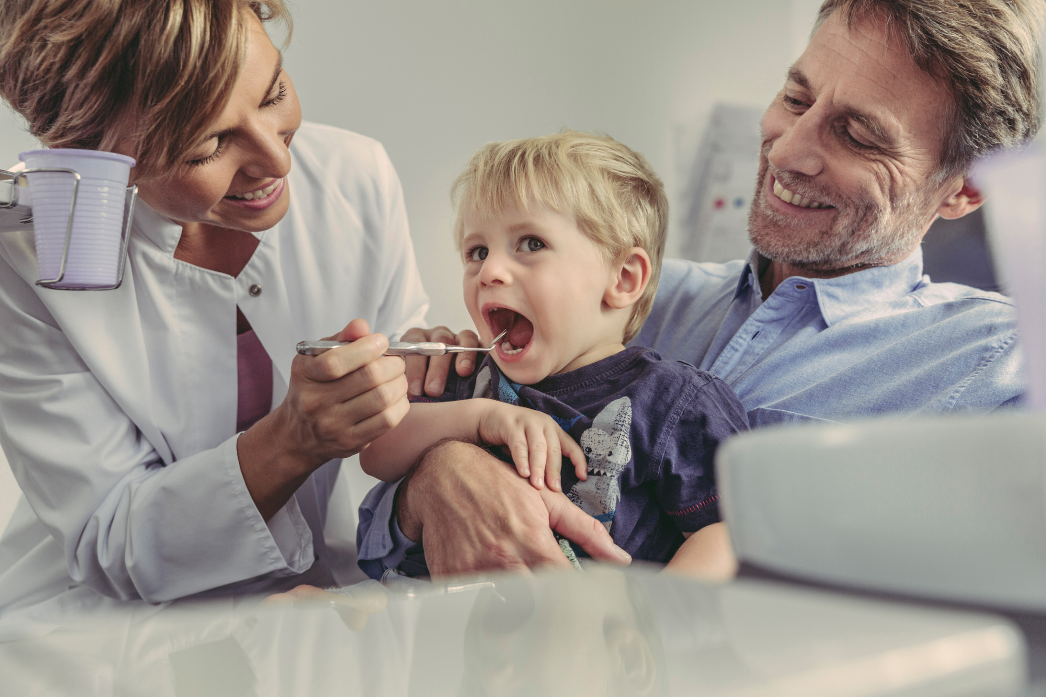 Female dentist looking into mouth of little boy sitting on father’s lap, Germany