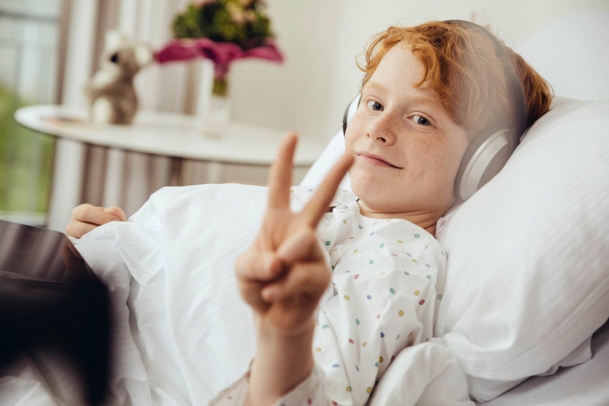 Young boy showing V sign while lying in hospital bed and listening to music