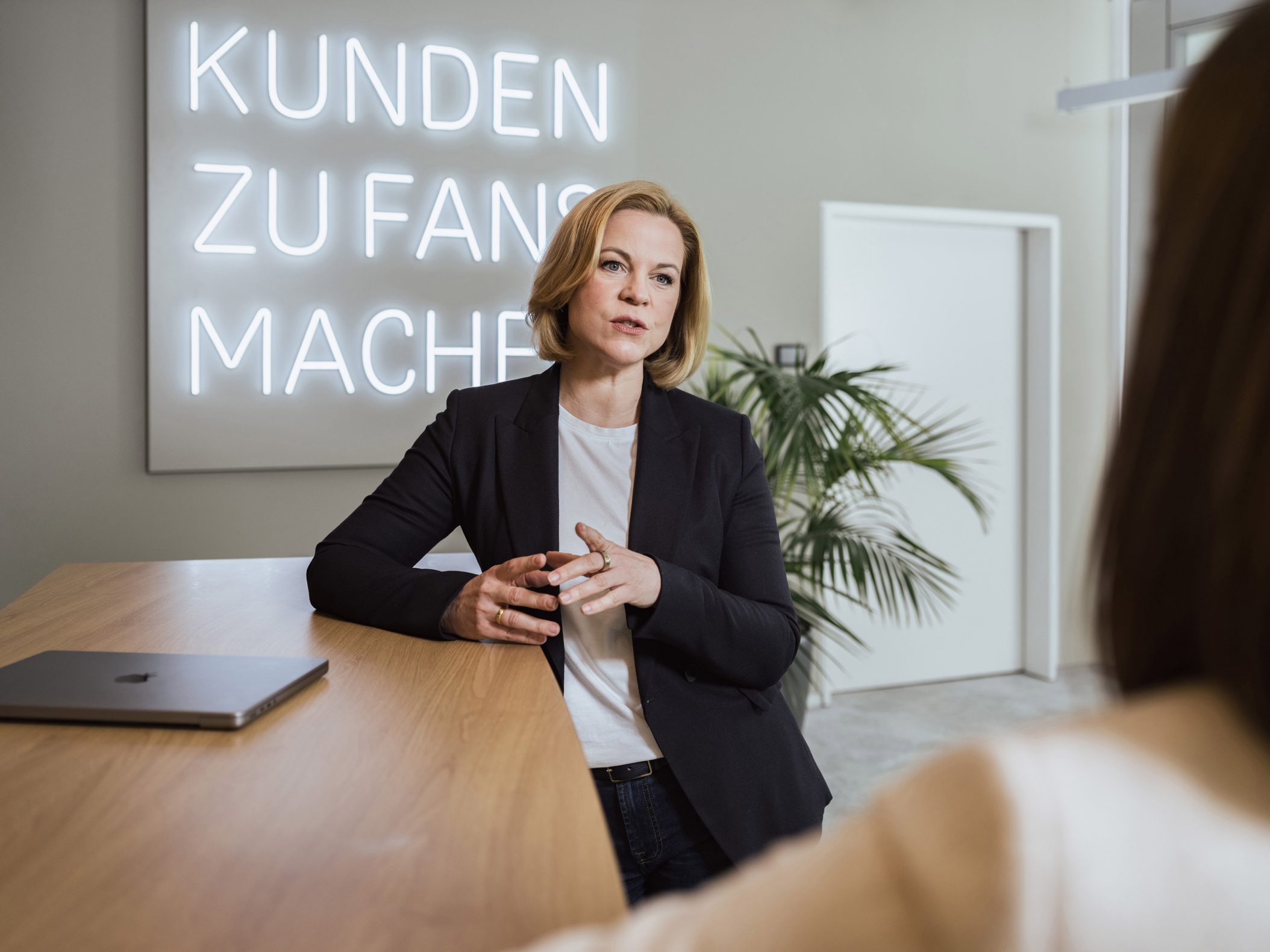Geschäftsfrau im Blazer spricht am Tisch vor Neonschrift