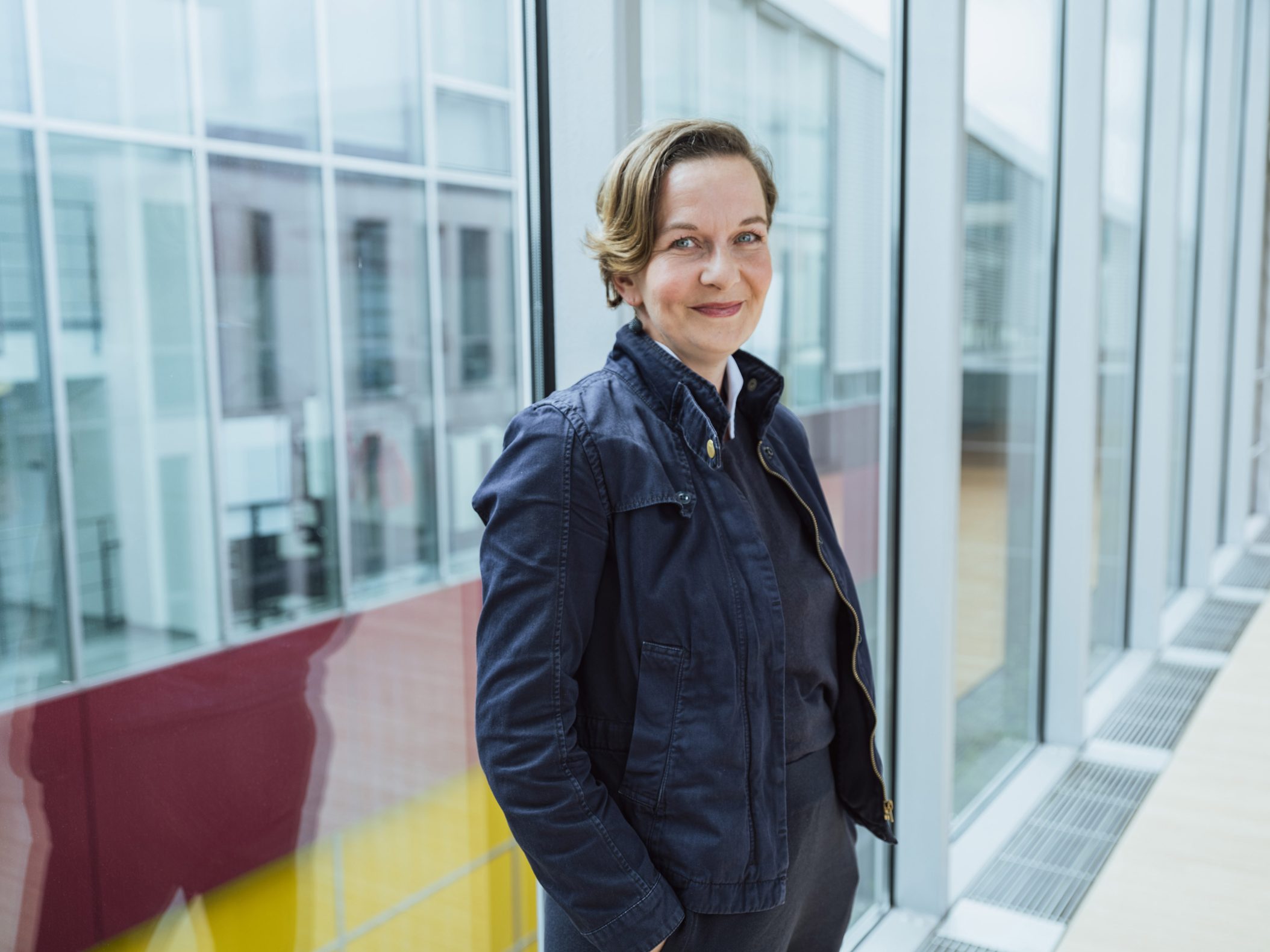 Lächelnde Frau in blauer Jacke vor modernem Glasgebäude