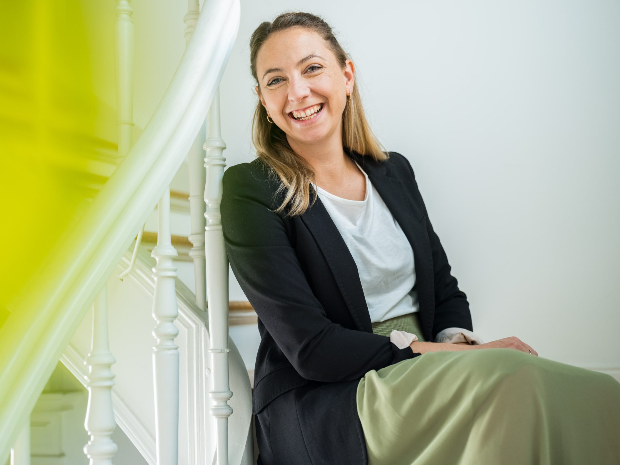 Smiling young woman sitting on white staircase in blazer by Mareen Fischinger