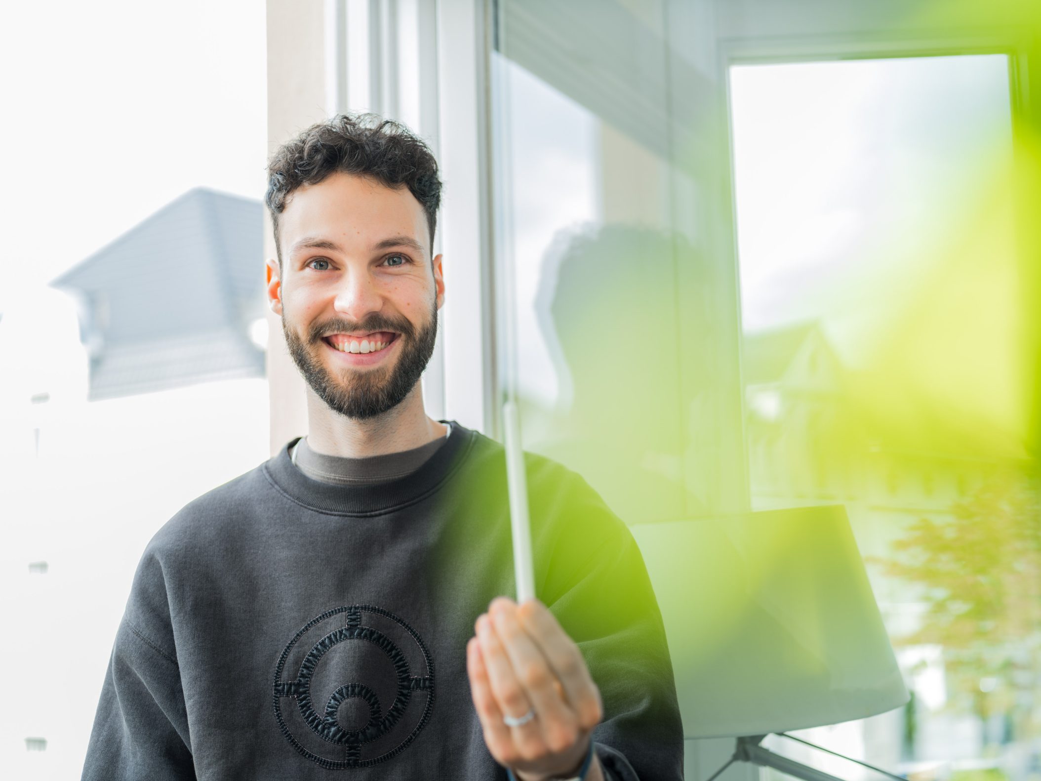 Smiling young bearded man holding transparent panel by window by Mareen Fischinger
